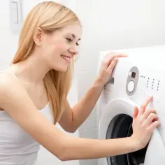 Uśmiechnięta blondynka przy pralce. Zastanawia się, dlaczego po użyciu zbyt dużej ilości proszku ubrania są lepkie i sztywne.