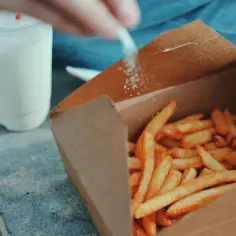 Ręka posypuje frytki solą. Obok stoi kubek z białym napojem. Sód dodaje smaku.