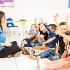 Dzieci na lekcji z podniesionymi rękami. Nauczycielka siedzi na podłodze. Frekwencja a ocena z zachowania - czy obecność wpływa na ocenę?