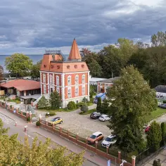 Czerwona willa z wieżyczką i restauracja z pomarańczowym dachem, gdzie można dobrze zjeść w Ustce. Wokół drzewa i parking z samochodami.