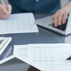 Osoba pracuje przy laptopie, analizując dokumenty i korzystając z kalkulatora. To może być związane z tym, czym jest umowa cywilnoprawna.