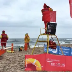 Ratownik wodny czuwa nad bezpieczeństwem na plaży. W oddali widać łódź ratowniczą i innych ratowników.