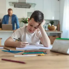 Chłopiec znużony odrabianiem lekcji, marzy o dniu bez prac domowych. W tle ojciec pracuje przy laptopie.