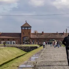 Para zwiedzająca Auschwitz indywidualnie idzie w stronę bramy obozu. W tle ceglane budynki i wieża strażnicza.