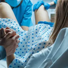 W szpitalu rodząca kobieta trzyma męża za rękę. Personel medyczny w niebieskich fartuchach asystuje. Czy balonik na wywołanie porodu ile trwa?