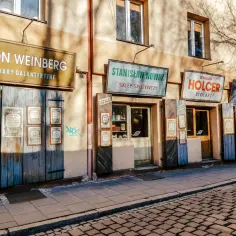 Stare szyldy sklepów na Podgórzu: Aron Weinberg (galanteria), Stanisław Nowak (spożywczy), Benjamin Holcer (stolarz), Salon Mód. Brukowana uliczka.