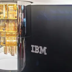 Złoty, wielopoziomowy rdzeń komputera kwantowego IBM, otoczony lustrzaną obudową, obok logo IBM.