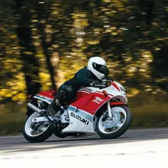 Motocyklista w kasku pędzi na czerwono-białym Suzuki RGV 250 przez leśną drogę.