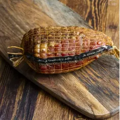 Wędzenie schabu na drewnianej desce. Widać siatkę, w którą jest owinięte mięso, oraz czarną wstążkę z napisem "...bo domowe jest lepsze!".