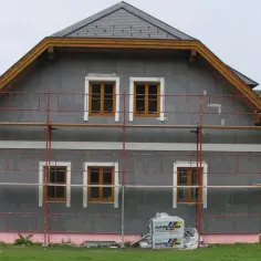 Dom w trakcie ocieplania. Szara fasada, drewniane okna, rusztowanie. Materiały budowlane Austrotherm wskazują na dbałość o niski współczynnik przewodzenia ciepła.