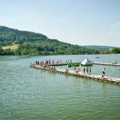 Ludzie odpoczywają na pomostach nad jeziorem, szukając ochłody gdzie nad wodę Dolny Śląsk. W tle zielone wzgórza.