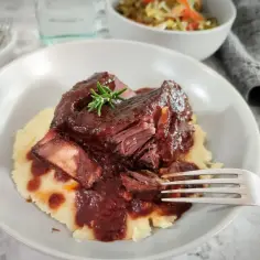 Pyszny szponder wołowy w sosie własnym, podany na purée ziemniaczanym, ozdobiony gałązką rozmarynu. Obok miseczka z surówką.