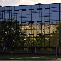 Nowoczesny budynek komercyjny z lustrzaną fasadą odbijającą zieleń drzew i pobliskie kamienice.