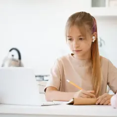 Dziewczynka z słuchawkami w uszach uczy się przy laptopie, notując w zeszycie. Tak wygląda nauka w chmurze.
