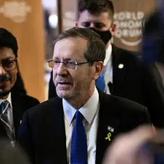 Prezydent Izraela, Isaac Herzog, w garniturze i z niebieskim krawatem, z przypinką z flagą Izraela i żółtą wstążką.