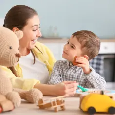 Uśmiechnięta opiekunka karmi chłopca. Na stole pluszowy miś i zabawki. Zastanawiasz się, ile opiekunki biorą za godzinę?