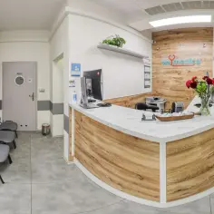 Recepcja kliniki medycznej z ladą, komputerem, kwiatami i poczekalnią.