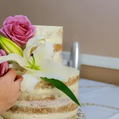 Ręce dekorują dwupiętrowy tort nagus, dodając róże i lilie. Piękny sposób, jak ozdobić tort kwiatami.
