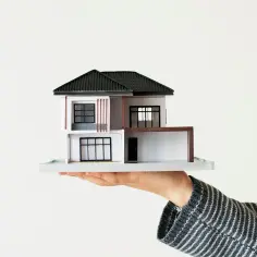 Dłoń trzyma miniaturowy obiekt budowlany – nowoczesny dom z dwuspadowym dachem i dużymi oknami.