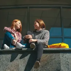 Dwie dziewczyny rozmawiają, siedząc na murku. Jedna z nich ma rude włosy i kratę. Czy to rozmowa o tym, czym jest absencja?