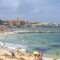 Tłum ludzi cieszy się słońcem i morzem na plaży w Tunezji, z widocznym w oddali zabytkowym fortem.