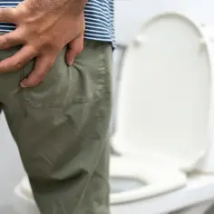 Mężczyzna trzyma się za pośladek, sugerując dyskomfort. W tle widać toaletę, co może wskazywać na problemy z wypróżnianiem, być może związane z owsikami w kale.