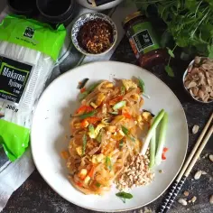Talerz z makaronem ryżowym w stylu Pad Thai, obok paczka makaronu, sos Pad Thai, orzeszki ziemne i chili.