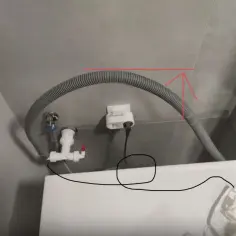 Szara rura odpływowa pralki w ścianie, podłączona do białego syfonu. Obok gniazdko elektryczne.