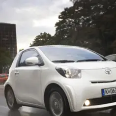Biały Toyota iQ jedzie po mokrej ulicy, mijając inne samochody. W tle widać budynek i drzewa.