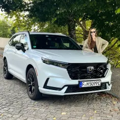 Uśmiechnięta kobieta opiera się o białą Honda CRV 2025 na brukowanej uliczce.
