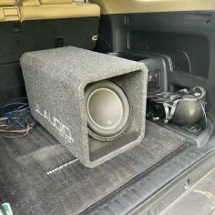 Szary subwoofer JL Audio w bagażniku samochodu, gotowy do montażu. Widać kable i akcesoria.