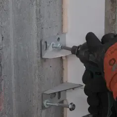 Rękawica z pomarańczowym wzmocnieniem przykręca metalowy wspornik do ściany. To część montażu, gdzie każdy element, jak ten kit, jest ważny.