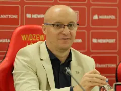 Nowy trener Widzewa w jasnej marynarce, siedzi na czerwonym fotelu z logo klubu.