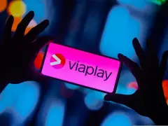 Dłonie trzymają smartfon z logo Viaplay. W tle rozmyte, kolorowe światła.
