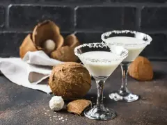 Dwa kieliszki z drinkiem Malibu z mlekiem, ozdobione wiórkami kokosowymi, obok świeżych kokosów i słodkich pralinek.