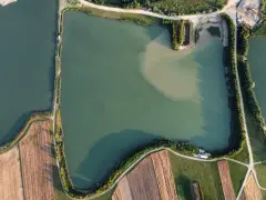 Widok z lotu ptaka na zielone oczko wodne, otoczone polami i drogami. Idealne miejsce na spokojne łowisko.
