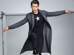 Joven con abrigo largo gris, camiseta negra con estrella blanca y vaqueros oscuros. Explora diversos tipos de abrigos para tu estilo.