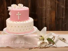 Elegancki dwupoziomowy tort na chrzest w różowo-białych barwach, ozdobiony krzyżem i kwiatami. Obok lilia, różaniec i świeca.