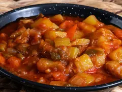 Un plato humeante de pisto manchego, con trozos de calabacín, pimiento y tomate en una salsa rojiza, servido en una cazuela negra.