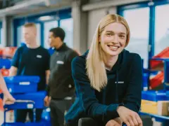 Lächelnde junge Frau im Lager, die ihren Werkstudentenjob genießt. Im Hintergrund arbeiten Kollegen mit blauen Behältern.