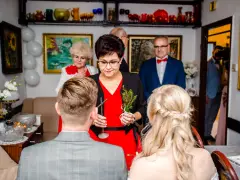 Rodzice błogosławią parę młodą. Kobieta w czerwonej sukience trzyma gałązkę, a obok stoi mężczyzna w garniturze.
