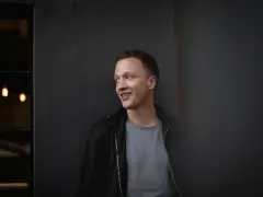 Joris, lächelnd, lehnt an einer dunklen Wand.
