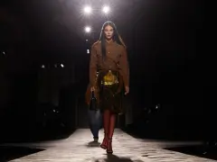 Modella su passerella, con gonna dorata e camicia marrone, un omaggio all'eleganza senza tempo di Prada, fondatore di un impero della moda.