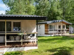 Dwa białe domki kempingowe w lesie, z werandami i meblami ogrodowymi. Idealne miejsce dla każdego **camping wagabunda**.