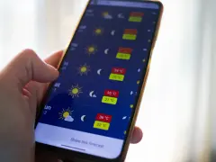 Dłoń trzyma smartfon z otwartą aplikacją pogoda, pokazującą prognozę z wysokimi temperaturami.