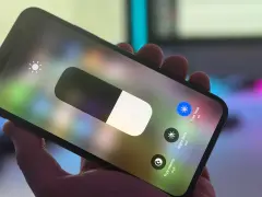 Dłoń trzyma iPhone'a z włączonym panelem sterowania. Widoczny suwak jasności i ikona True Tone.