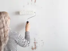Mujer con cabello rubio y camiseta a rayas está a punto de empapelar pared con rodillo blanco.