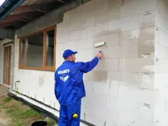 Pracownik w niebieskim kombinezonie nakłada wałkiem grunt na ścianę z bloczków. To kluczowy etap przygotowania pod styropian.