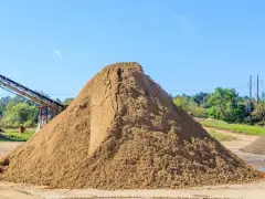 Ogromna hałda piasku na placu budowy, zastanawiasz się, ile kosztuje tona piasku? W tle taśmociąg i drzewa.