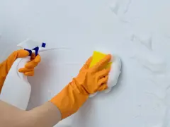 Manos con guantes naranjas rociando y limpiando paredes con espuma y esponja.